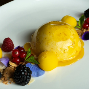 A delicious mango mousse surrounded by fresh berries and edible flowers on a white plate.